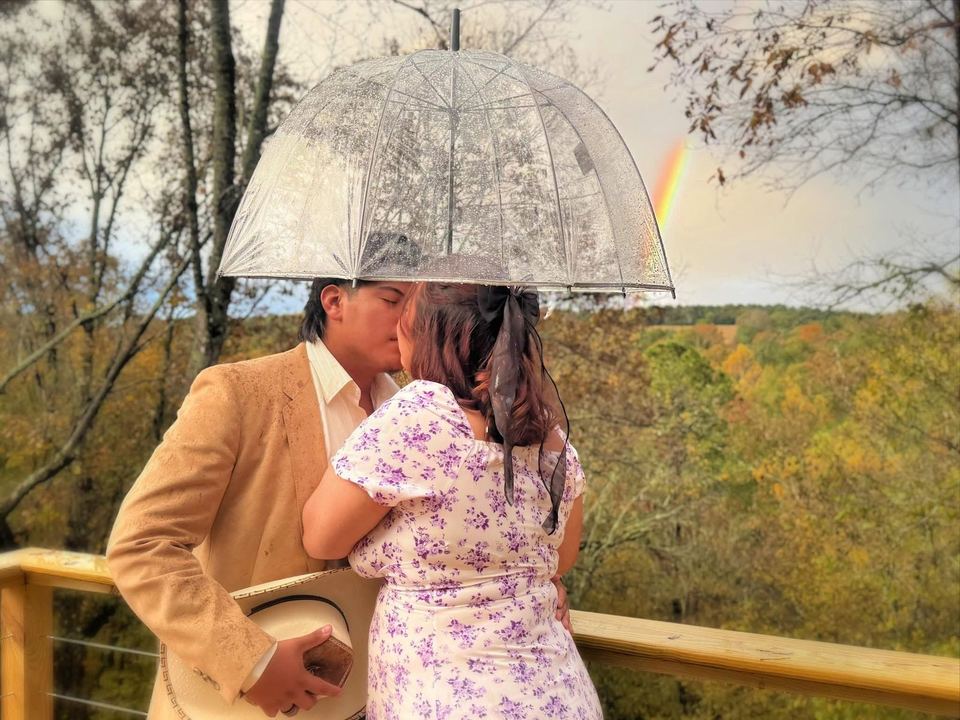 A couple kissing under a clear umbrella on the deck, overlooking the fall valley