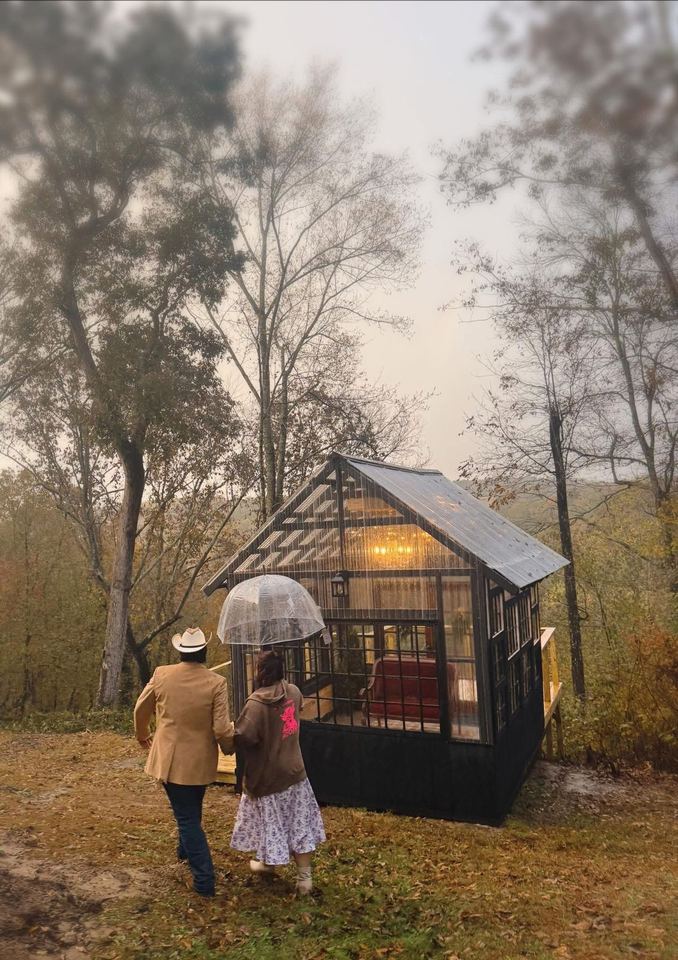 A couple with a clear umbrella walking toward the glowing Glasshouse in the woods